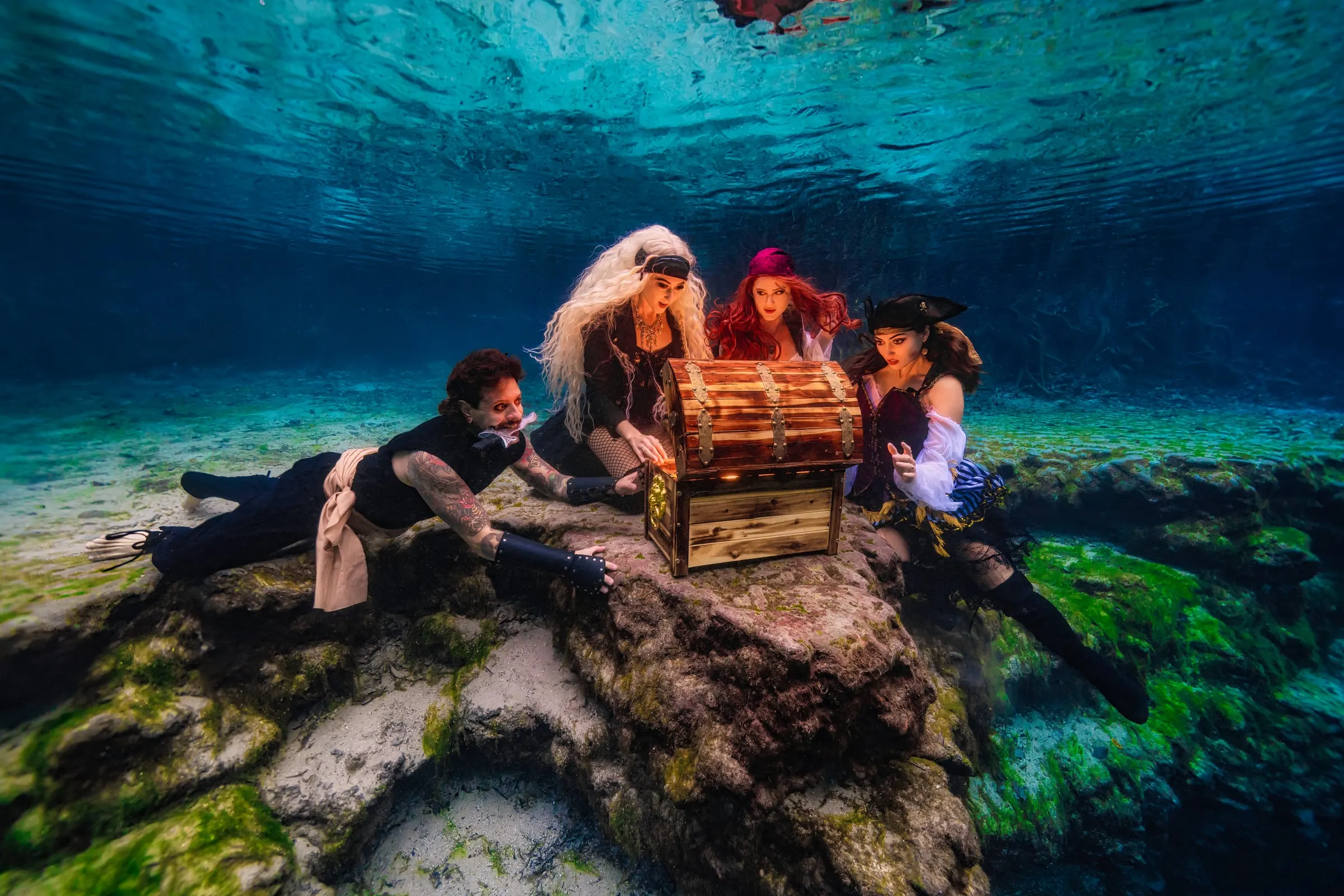 Four costumed pirates crowd around a glowing treasure chest on a mossy rock in a clear cenote, light rippling on the surface above them.