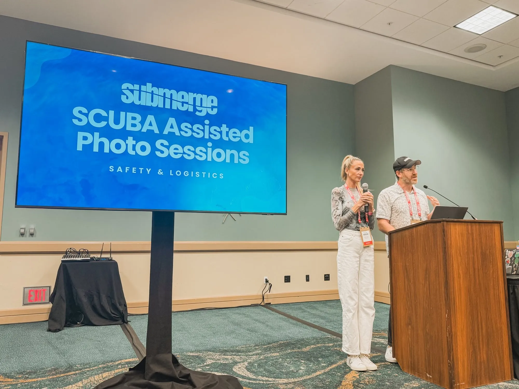 Abbey and Justin on stage at a dive-industry conference, a slide behind them titled SCUBA Assisted Photo Sessions: Safety and Logistics.