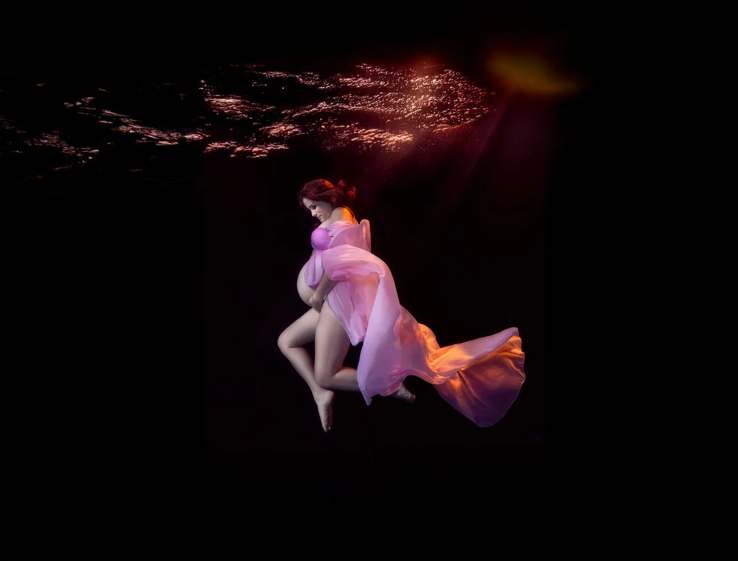 An expectant mother in pink and lavender fabric floats in near-black water, warm light catching the trailing cloth.